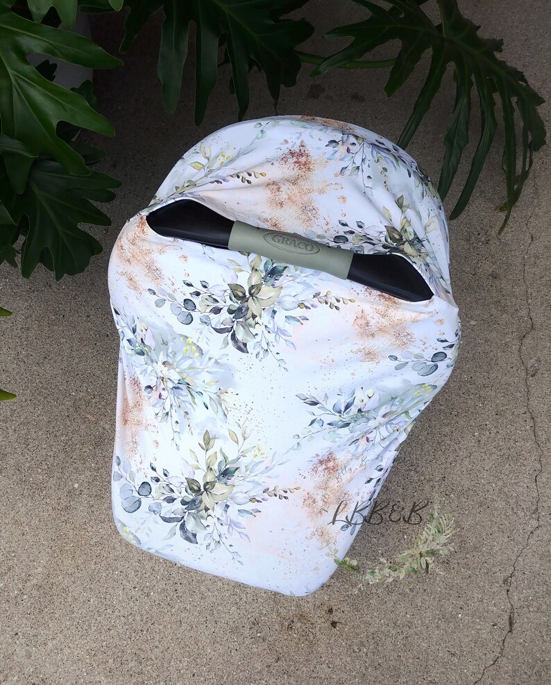 Puede incluir: Una funda blanca para asiento de coche de beb&eacute; con un dise&ntilde;o floral en tonos verdes, grises y dorados. La funda tiene un asa Graco y est&aacute; colocada sobre una superficie de hormig&oacute;n. El patr&oacute;n floral incluye hojas y ramas.