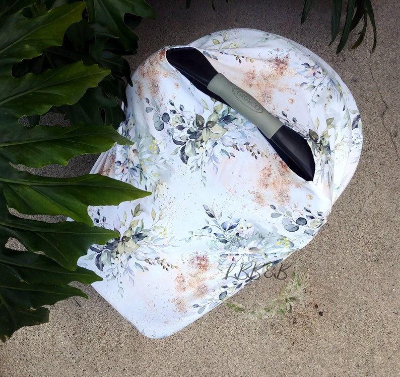 Puede incluir: Una funda blanca para asiento de coche de beb&eacute; con un dise&ntilde;o floral con hojas verdes y azules y detalles dorados. La funda est&aacute; colocada sobre un asa de asiento de coche negro y gris. La marca "Graco" es visible.