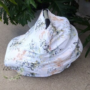 Puede incluir: Una funda blanca para asiento de coche de beb&eacute; con un estampado floral. La funda presenta un dise&ntilde;o de hojas verdes, detalles dorados y detalles en azul claro. Las letras "LBB&B" son visibles en la parte inferior izquierda.