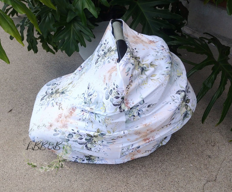 Puede incluir: Una funda blanca para asiento de coche de beb&eacute; con un estampado floral. La funda presenta un dise&ntilde;o de hojas verdes, detalles dorados y detalles en azul claro. Las letras "LBB&B" son visibles en la parte inferior izquierda.