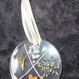 May include: A round, clear glass ornament with a white ribbon. The ornament is divided into four sections, each containing a different element: dried lavender, white crystals, a sprig of rosemary, and a copper heart.