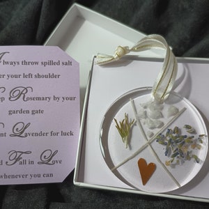 May include: A clear round ornament with a white background and four sections. Each section contains a different element: a sprig of rosemary, a heart, white crystals, and lavender flowers. The ornament is hanging from a gold ribbon and is in a white box with a purple card that reads "Always throw spilled salt over your left shoulder Keep Rosemary by your garden gate Plant Lavender for luck And Fall in Love whenever you can."