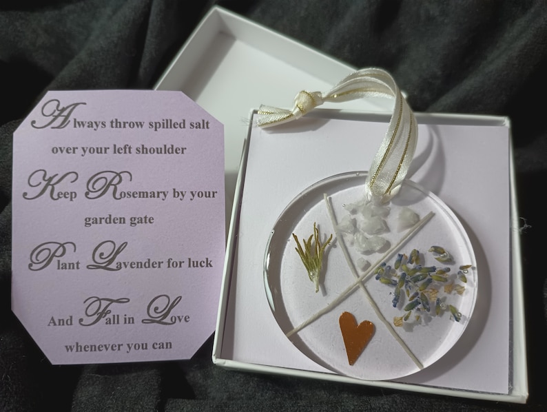 May include: A clear round ornament with a white background and four sections. Each section contains a different element: a sprig of rosemary, a heart, white crystals, and lavender flowers. The ornament is hanging from a gold ribbon and is in a white box with a purple card that reads "Always throw spilled salt over your left shoulder Keep Rosemary by your garden gate Plant Lavender for luck And Fall in Love whenever you can."