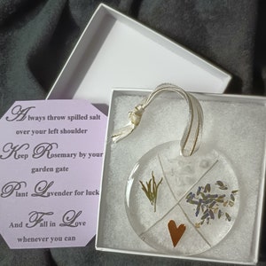 May include: A white box with a round, clear glass ornament inside. The ornament has a gold ribbon and a heart shape in the center. The ornament is filled with dried rosemary, lavender, and a heart. The box has a purple card with the text: "Always throw spilled salt over your left shoulder. Keep Rosemary by your garden gate. Plant Lavender for luck. And Fall in Love whenever you can."