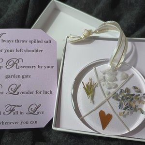 May include: A round, clear resin ornament with a copper heart, dried rosemary, lavender, and white crystals. The ornament is in a white box with a tag that reads: "Always throw spilled salt over your left shoulder. Keep Rosemary by your garden gate. Plant Lavender for luck. And Fall in Love whenever you can."