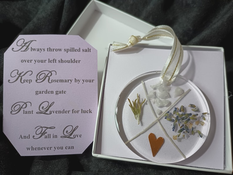 May include: A round, clear resin ornament with a copper heart, dried rosemary, lavender, and white crystals. The ornament is in a white box with a tag that reads: "Always throw spilled salt over your left shoulder. Keep Rosemary by your garden gate. Plant Lavender for luck. And Fall in Love whenever you can."