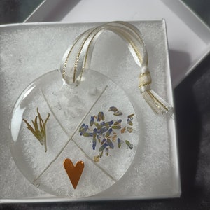 May include: A round glass ornament with a gold ribbon. The ornament is divided into four sections, each containing a different type of dried flower. The center section features a small, copper-colored heart.