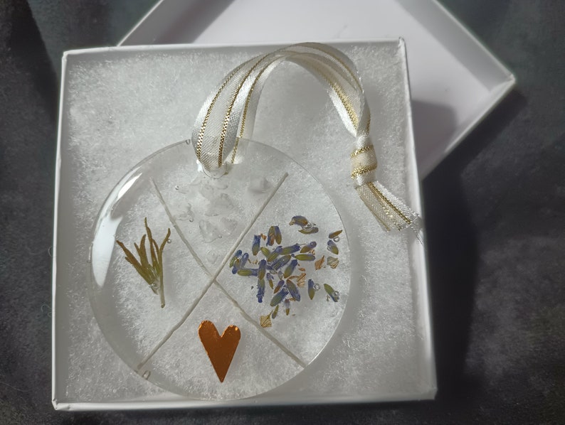 May include: A round glass ornament with a gold ribbon. The ornament is divided into four sections, each containing a different type of dried flower. The center section features a small, copper-colored heart.