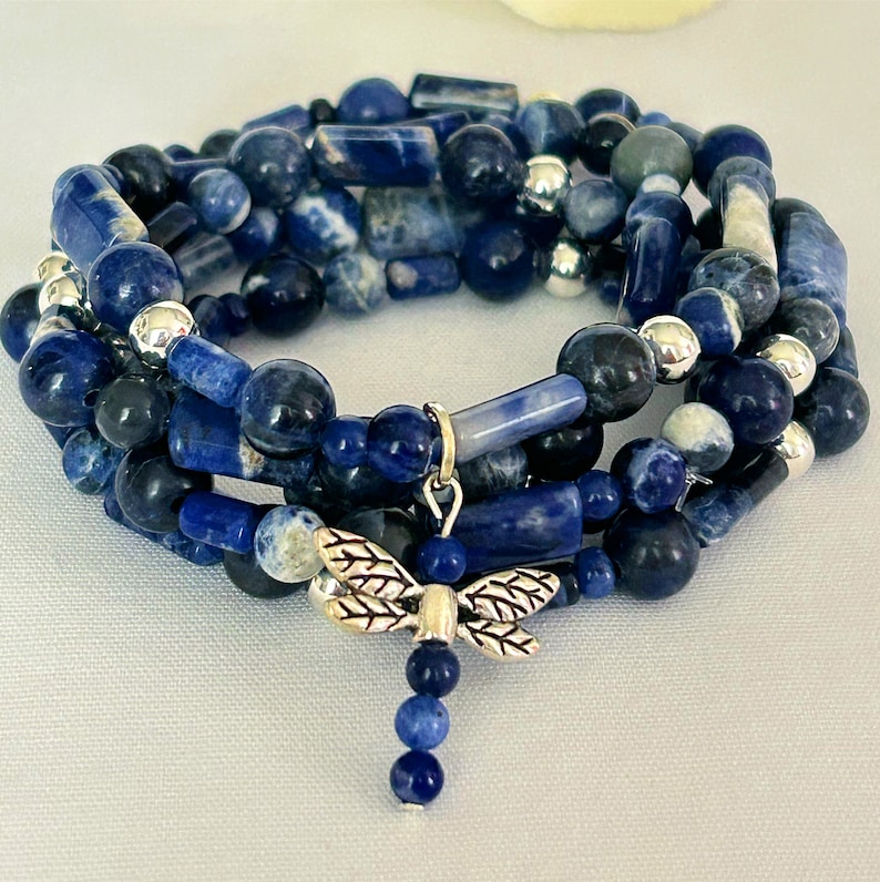 May include: A wrap bracelet featuring blue sodalite beads in various shapes and sizes, accented with silver-colored beads. A silver dragonfly charm dangles from the bracelet, adding a decorative touch. The bracelet is designed to wrap multiple times around the wrist.