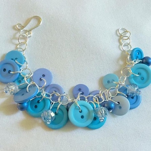 May include: A handmade bracelet featuring a collection of blue buttons in various shades, including light, medium, and dark blues. The buttons are connected by a silver chain and accented with small crystal beads. The bracelet has a hook clasp.