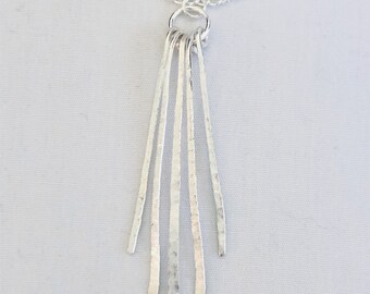May include: A silver-colored necklace with a chain and a pendant featuring five vertical, textured bars of varying lengths. The bars are attached to a small ring, which is connected to the chain. The necklace is displayed against a white background.