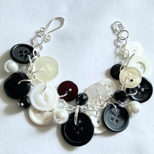 May include: A charm bracelet constructed from silver-toned metal links featuring an assortment of black, white, and translucent buttons of varying sizes, interspersed with small black and white spherical beads, finished with a hook clasp.
