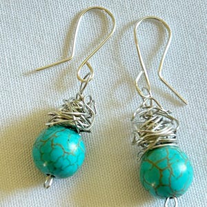 May include: A pair of turquoise bead earrings with silver wire wrapping and silver ear hooks. The turquoise beads have a brown vein pattern. The earrings are displayed on a white fabric background.