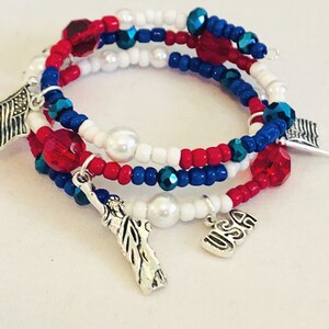 May include: A patriotic wrap bracelet featuring red, white, and blue beads with silver charms. The bracelet includes charms of the American flag, the Statue of Liberty, and the letters "USA". The beads are a mix of small round beads and larger faceted beads.