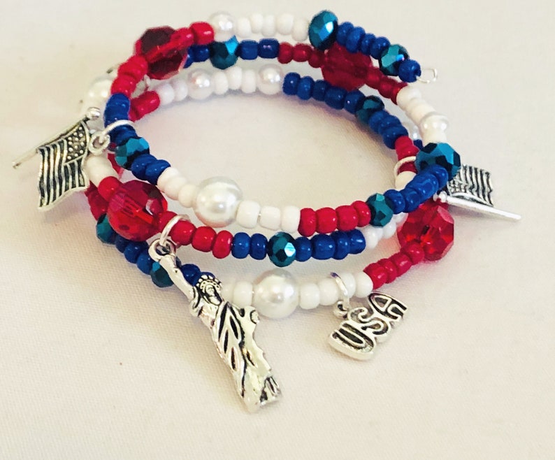 May include: A patriotic wrap bracelet featuring red, white, and blue beads with silver charms. The bracelet includes charms of the American flag, the Statue of Liberty, and the letters "USA". The beads are a mix of small round beads and larger faceted beads.