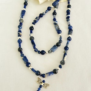 May include: A necklace and bracelet set featuring blue sodalite beads and silver accents. The necklace has a dragonfly charm. The beads are a mix of round and rectangular shapes. The jewelry is displayed on a white surface.