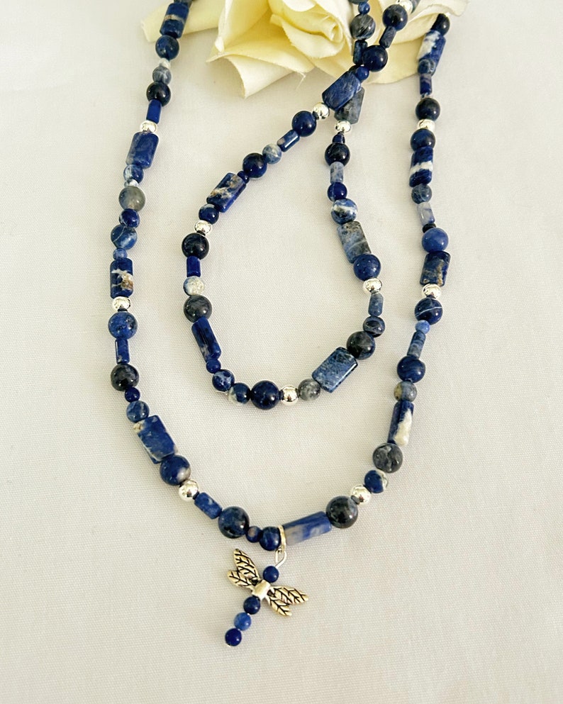 May include: A necklace and bracelet set featuring blue sodalite beads and silver accents. The necklace has a dragonfly charm. The beads are a mix of round and rectangular shapes. The jewelry is displayed on a white surface.