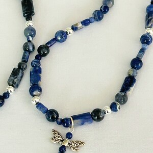 May include: A beaded necklace featuring blue sodalite stones in various shapes and sizes, with silver accents. A silver dragonfly charm hangs from the center of the necklace. The necklace is displayed on a white surface.