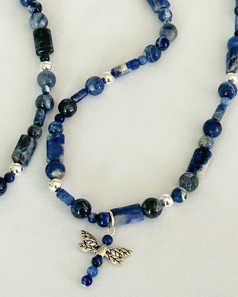 May include: A beaded necklace featuring blue sodalite stones in various shapes and sizes, with silver accents. A silver dragonfly charm hangs from the center of the necklace. The necklace is displayed on a white surface.