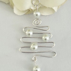 May include: A silver-toned pendant necklace featuring a unique, abstract design. The necklace has a series of horizontal lines with faux pearl accents, and a spiral element. The necklace is displayed on a white background, suggesting a jewelry theme.