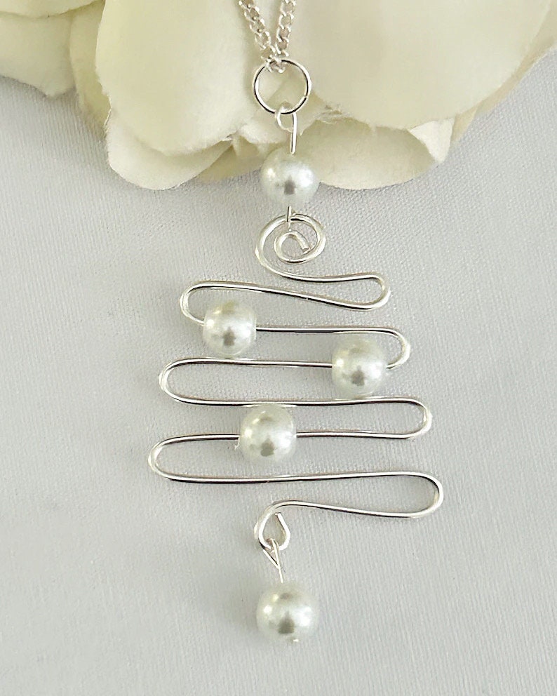 May include: A silver-toned pendant necklace featuring a unique, abstract design. The necklace has a series of horizontal lines with faux pearl accents, and a spiral element. The necklace is displayed on a white background, suggesting a jewelry theme.