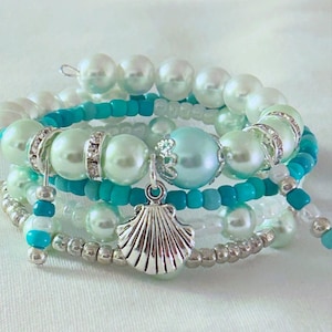 May include: A multi-strand wrap bracelet featuring a combination of white and light blue faux pearls, turquoise seed beads, and small silver-toned metallic beads. A silver-toned seashell charm hangs from the center, accented by small clear crystals.