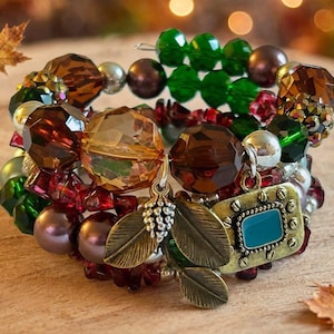 May include: A multi-strand wrap bracelet featuring a mix of faceted brown, green, and red beads, smooth metallic spheres, and small silver-toned charms. The charms include two metal leaves and a rectangular frame with a central teal inlay.