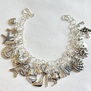 Pode incluir: Pulseira de berloques prateada com v&aacute;rios berloques, incluindo uma estrela do mar, um salto alto, um biqu&iacute;ni e uma concha. A pulseira tem um fecho de lagosta e &eacute; feita de correntes de prata interligadas. Os berloques s&atilde;o detalhados e adicionam um toque decorativo.