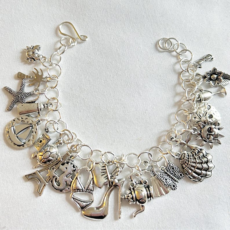 Pode incluir: Pulseira de berloques prateada com v&aacute;rios berloques, incluindo uma estrela do mar, um salto alto, um biqu&iacute;ni, uma concha e uma tartaruga. A pulseira tem um fecho de lagosta e &eacute; exibida em uma superf&iacute;cie branca. Os berloques detalhados adicionam um toque &uacute;nico.