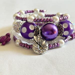 May include: A stack of beaded bracelets in shades of purple, white, and silver. The bracelets feature various bead sizes and shapes, including large purple polka-dot beads, pearl-like beads, and a silver charm. A purple turtle charm is visible.
