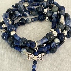 May include: A multi-strand bracelet featuring blue sodalite beads in various shapes and sizes, interspersed with silver-colored beads. A silver dragonfly charm dangles from the bracelet, adding a decorative touch. The bracelet is designed to be wrapped around the wrist.