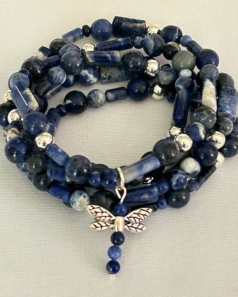 May include: A multi-strand bracelet featuring blue sodalite beads in various shapes and sizes, interspersed with silver-colored beads. A silver dragonfly charm dangles from the bracelet, adding a decorative touch. The bracelet is designed to be wrapped around the wrist.