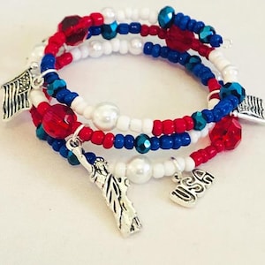May include: A patriotic wrap bracelet featuring red, white, and blue beads. The bracelet includes charms of the American flag, the Statue of Liberty, and the letters "USA". The beads are a mix of small round beads and larger faceted beads.