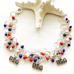 May include: A silver chain bracelet with red, white, and blue beads and three "USA" charms. The bracelet is displayed on a white surface with a starfish in the background. The bracelet is a patriotic accessory.