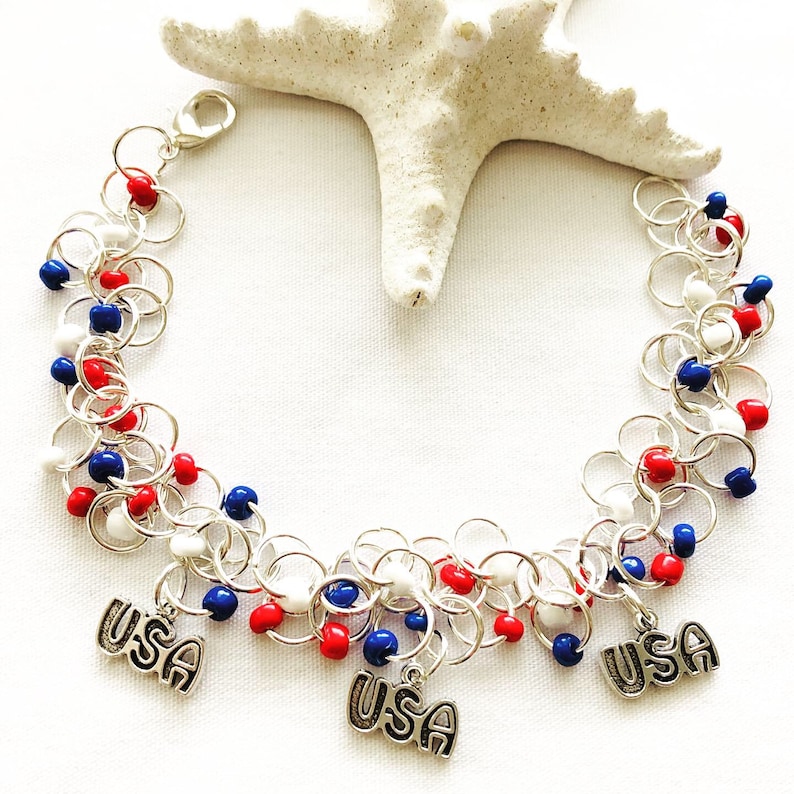 May include: A silver chain bracelet with red, white, and blue beads and three "USA" charms. The bracelet is displayed on a white surface with a starfish in the background. The bracelet is a patriotic accessory.