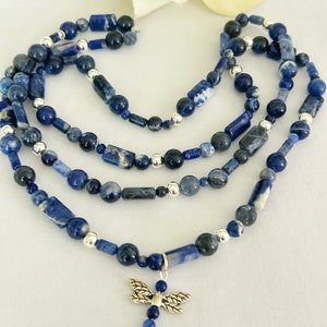 May include: A layered necklace featuring blue sodalite beads in various shapes and sizes, interspersed with silver-colored beads. A silver dragonfly charm hangs from the bottom layer. The necklace is displayed on a white surface.