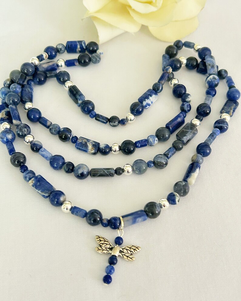May include: A layered necklace featuring blue sodalite beads in various shapes and sizes, interspersed with silver-colored beads. A silver dragonfly charm hangs from the bottom layer. The necklace is displayed on a white surface.