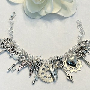 May include: Silver charm bracelet featuring various charms, including keys, wings, a heart, a gear, and a bird. The bracelet is made of linked chain and has a lobster clasp closure. The charms are detailed and have an antique appearance.