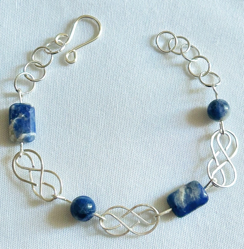 May include: A silver bracelet featuring blue lapis lazuli beads and rectangular stones. The bracelet incorporates infinity symbols and chain links. The clasp is a simple hook and eye closure. The bracelet is displayed on a light blue surface.