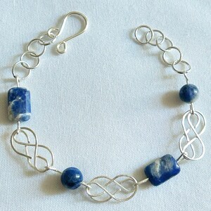 May include: A silver bracelet featuring blue lapis lazuli beads and rectangular stones. The bracelet incorporates infinity symbols and chain links. The clasp is a simple hook and eye closure. The bracelet is displayed on a light blue surface.