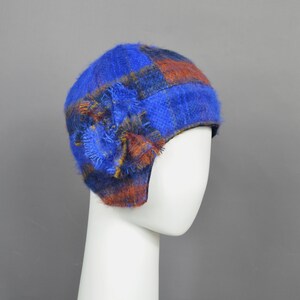 May include: A blue and orange plaid cloche hat with a decorative flower on the side. The hat is made of a fuzzy material and is displayed on a white mannequin head. The hat is a winter accessory.