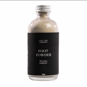 May include: A glass jar with a black label that reads "Dirty Dude Apothecary Foot Powder Dirty shoes, healthy feet 4oz". The jar is filled with a light brown powder.
