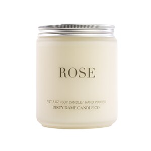 May include: A white candle jar with a silver lid. The jar is labeled "ROSE" and "NET 8 OZ/SOY CANDLE/ HAND POURED DIRTY DAME CANDLE CO."