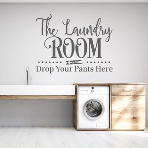 Op de afbeelding: Een witte wasruimte met een witte wasmachine, een houten aanrecht en een witte gootsteen. De muur heeft een grijze vinyl sticker met de tekst "The Laundry Room Drop Your Pants Here" met een wasknijper afbeelding.