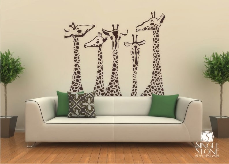 May include: A white couch with two green and one brown patterned pillows in front of a wall with a brown giraffe family silhouette decal.