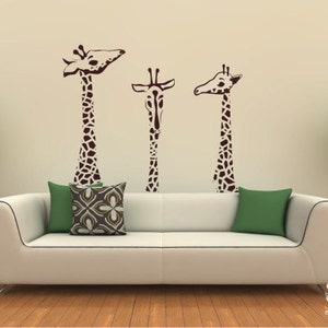 May include: Three brown giraffe silhouettes are on a beige wall above a white couch with green and brown patterned throw pillows. The couch is in front of a wood floor.
