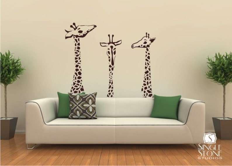 May include: Three brown giraffe silhouettes are on a beige wall above a white couch with green and brown patterned throw pillows. The couch is in front of a wood floor.