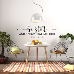 May include: A black and white wall decal with the text "be still and know that I am God Psalm 46:10". The decal is displayed on a white wall in a dining room setting with a table, chairs, and plants.