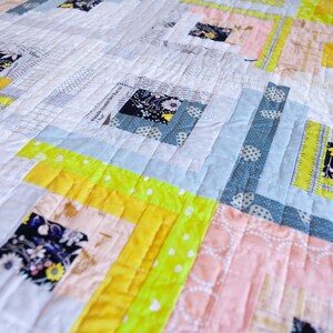 May include: A quilt with a patchwork design featuring squares of different fabrics in shades of yellow, pink, blue, and white. The quilt is quilted with a straight stitch pattern.