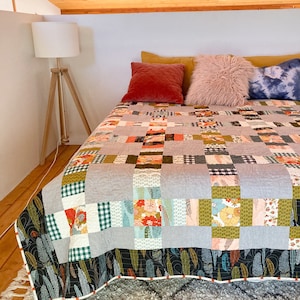May include: A gray and white patchwork quilt with a floral and geometric pattern. The quilt is on a bed with a variety of pillows, including a pink faux fur pillow and a blue tie-dye pillow.
